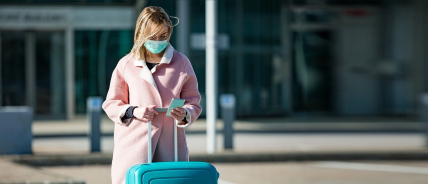 Ratgeber Corona und Reiserecht - Frau mit Schutzmaske und Koffer guckt auf Smartphone